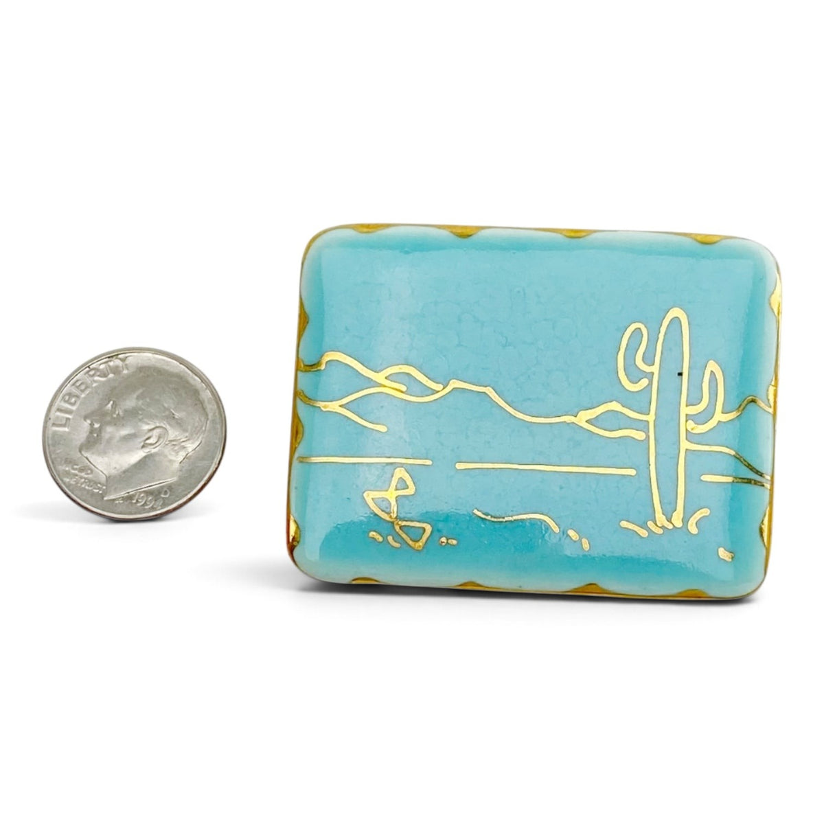 FuzzBeez turquoise ceramic brooch with gold saguaro cactus line art and scalloped edges next to a US dime for scale